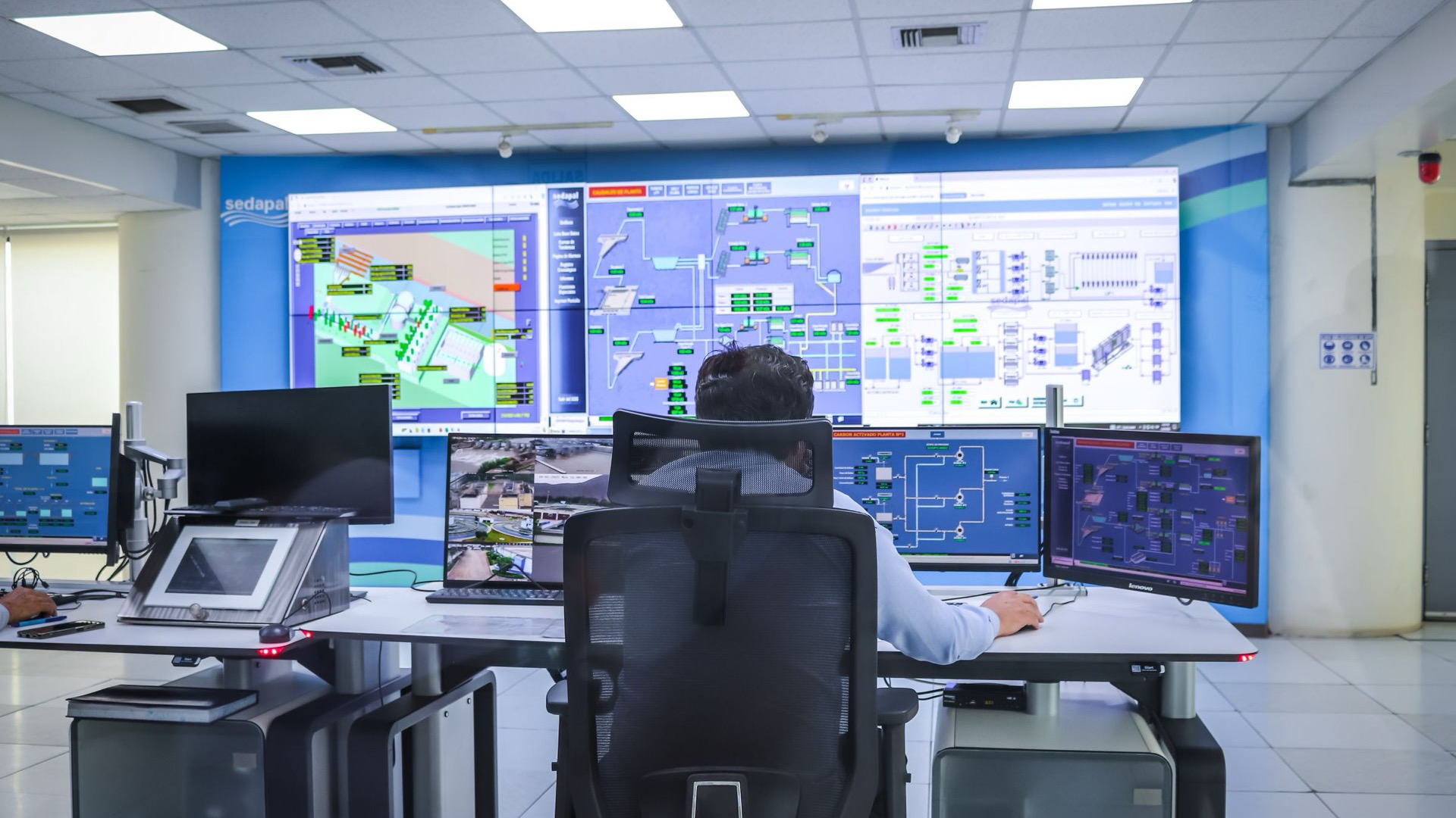 Sala de control industrial con esquemas SCADA en pantalla y un operador en su puesto